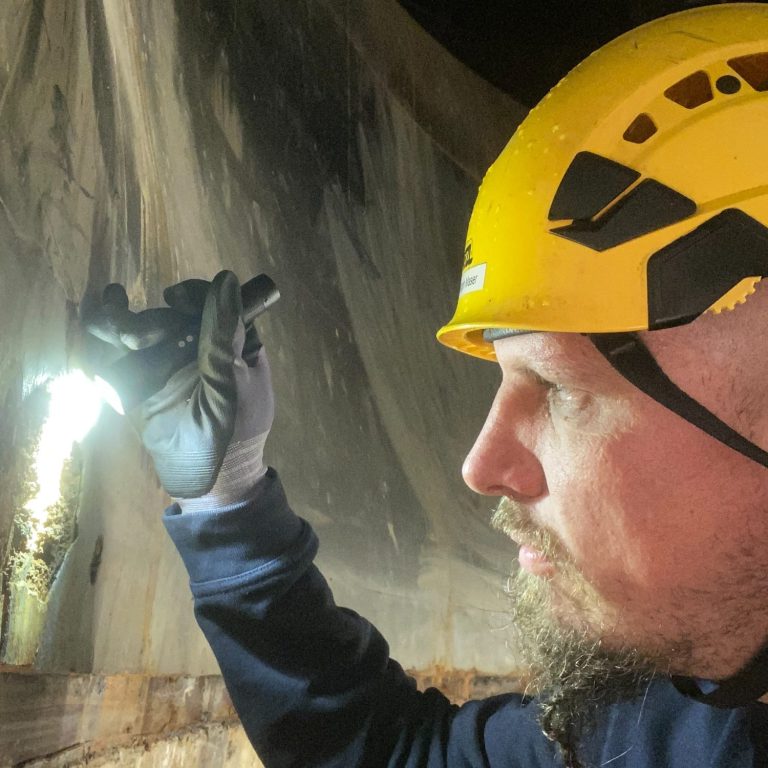 Christoph Maser | Der Korrosionsschützer Mann mit Helm und Sicherheitsjacke, lächelt vor grünem Hintergrund.